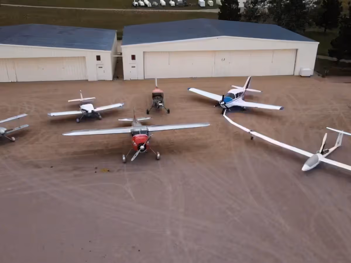Gliders and light aircraft parked on the ramp at Kelly Air Park beside two hangars.