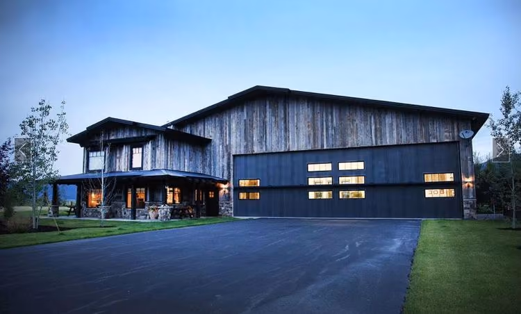 A barnwood hangar home at dusk with the interior lit through the hangar door windows.