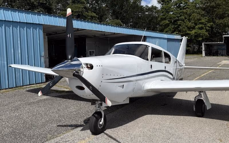 A Piper Comanche parked at the open door of a blue steel hangar.
