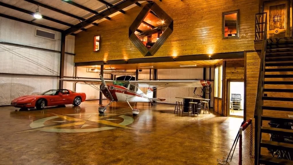 A hangar interior finished as living space: wood panelling, a mezzanine window, a light aircraft and a sports car on stained concrete.