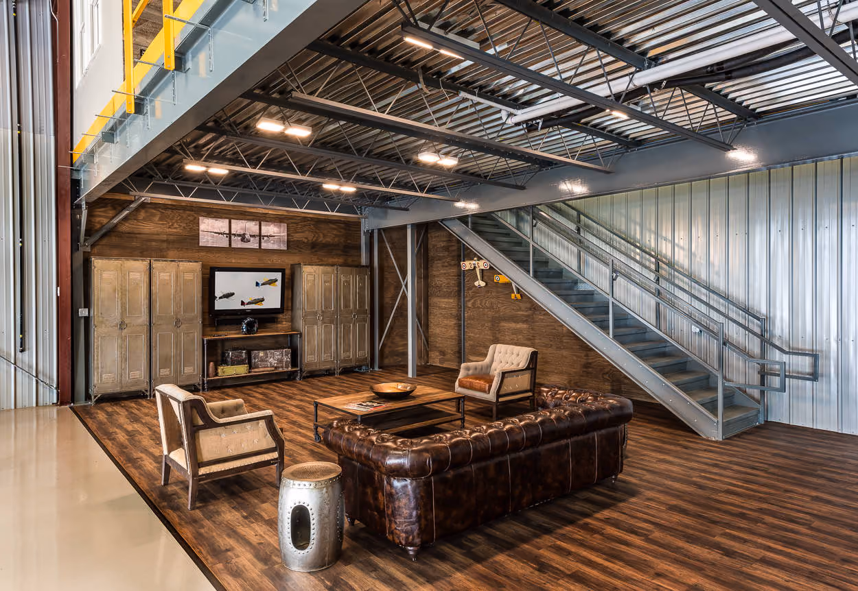 A hangar lounge under a steel mezzanine: leather sofa, lockers and a staircase beside the aircraft bay.