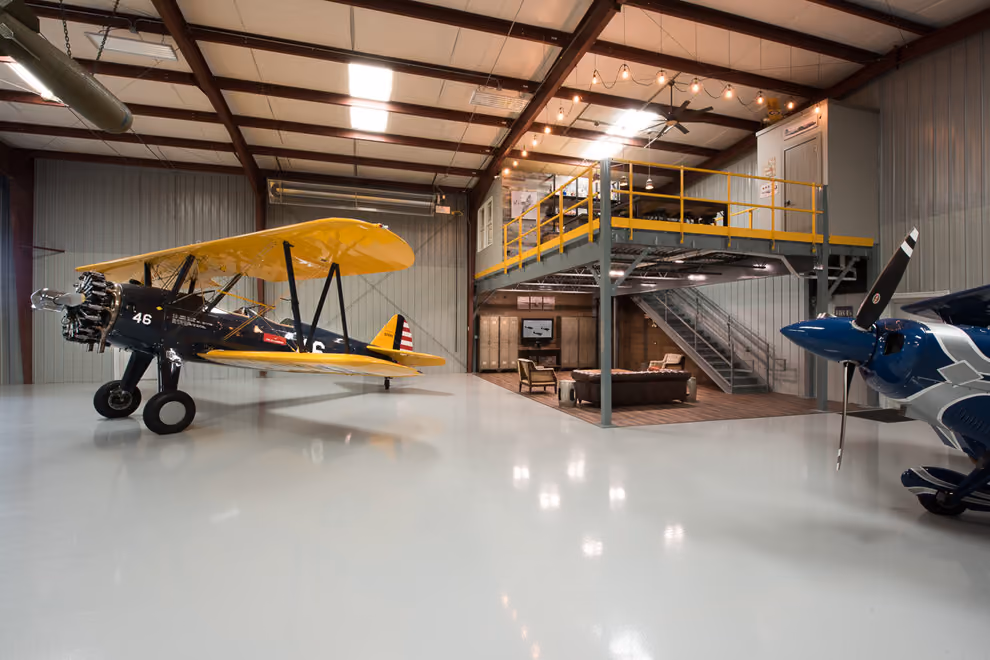 A staged hangar lounge with a chesterfield sofa beneath a steel mezzanine.