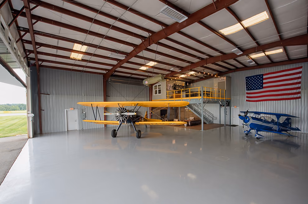 A yellow biplane and a blue aerobatic aircraft in a steel hangar with a mezzanine office.