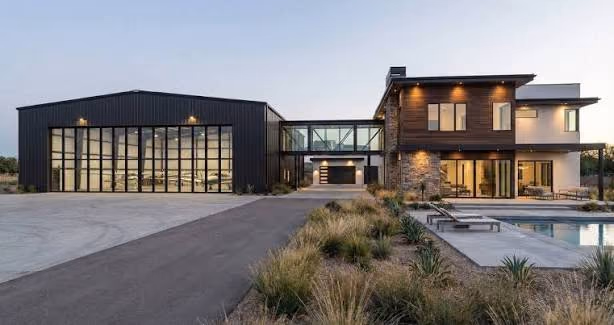 A modern black steel hangar joined to a two-storey home by a glass connector, with a pool in the foreground.
