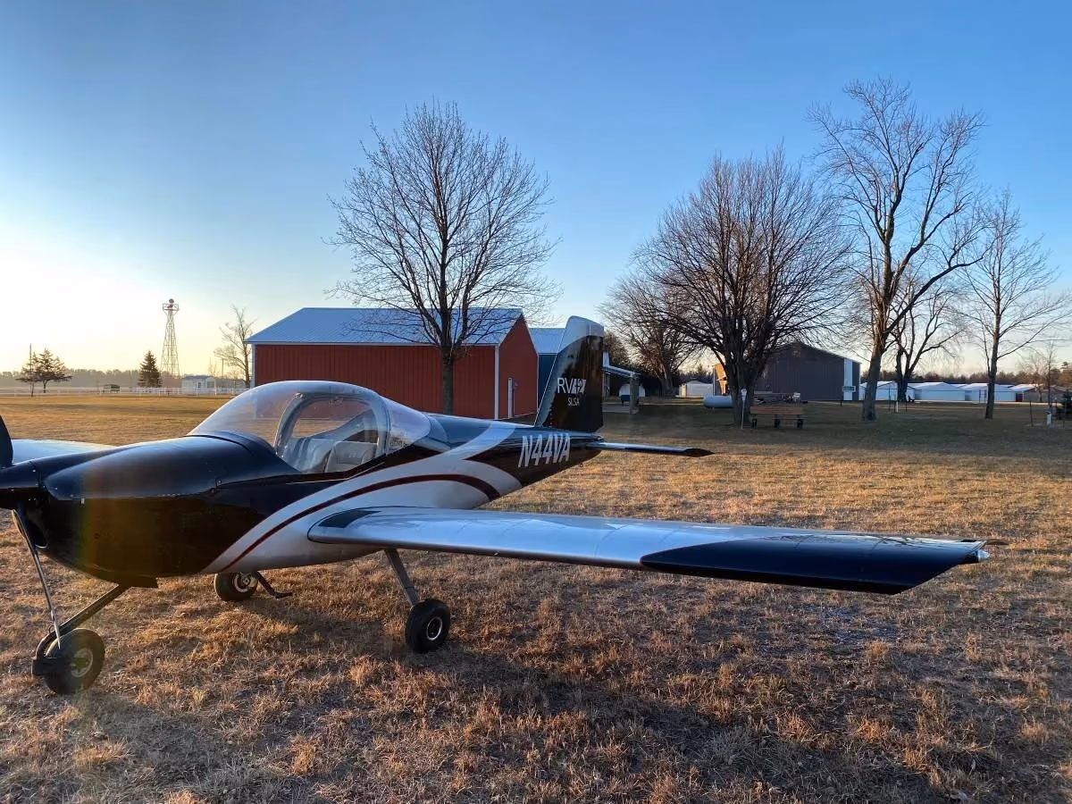 A light sport aircraft parked on winter grass beside a red barn hangar.