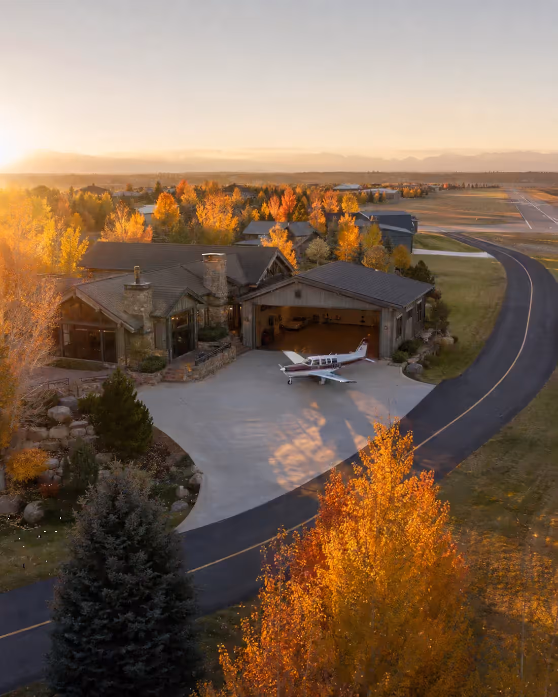 An airpark residence from the air at sunrise, its aircraft on the apron and the taxiway curving out to the runway