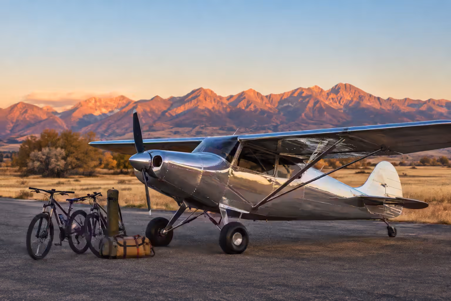 A polished aluminium taildragger at first light with two mountain bikes and duffels staged beside the wing, alpenglow on the range behind