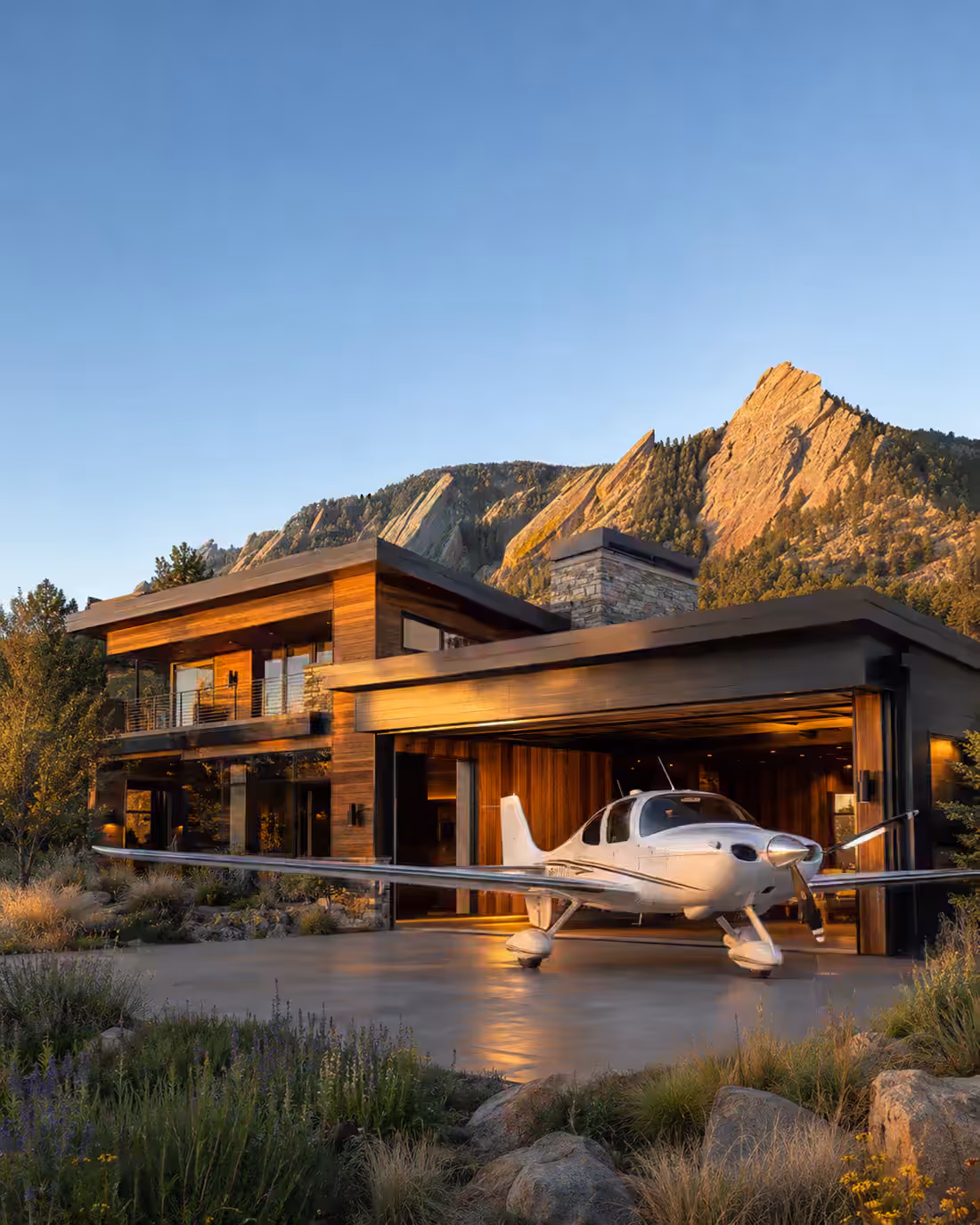 A single-engine aircraft parked on the apron outside the open hangar bay of a cedar and stone residence at golden hour