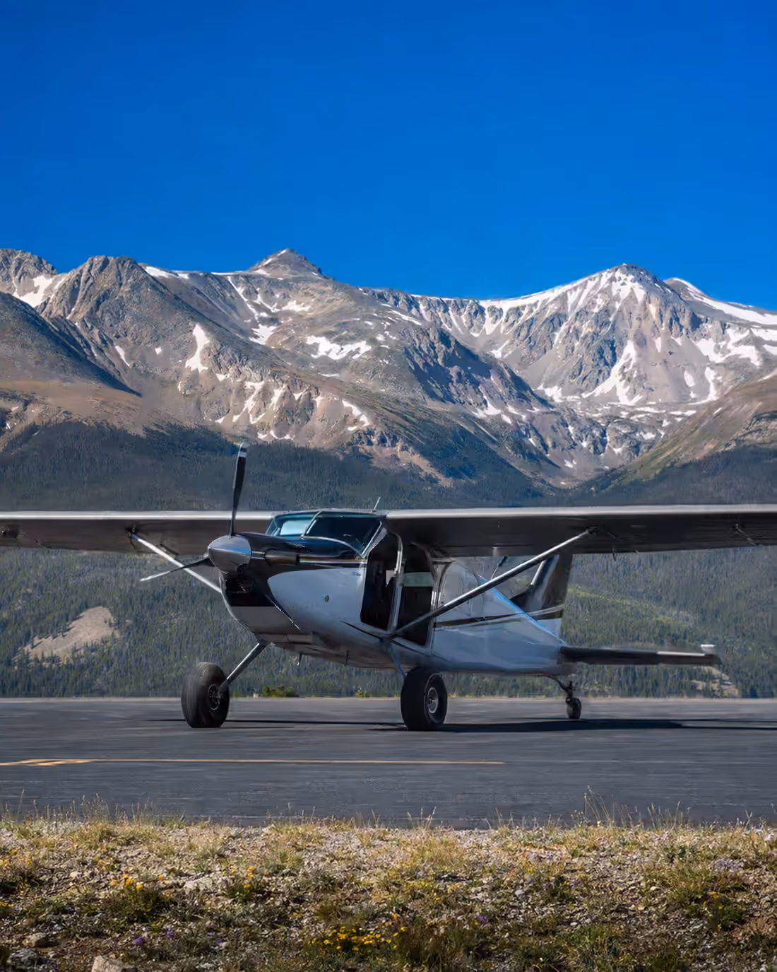 A high-wing taildragger on a high-country ramp with its cabin door open, a snow-streaked ridge behind
