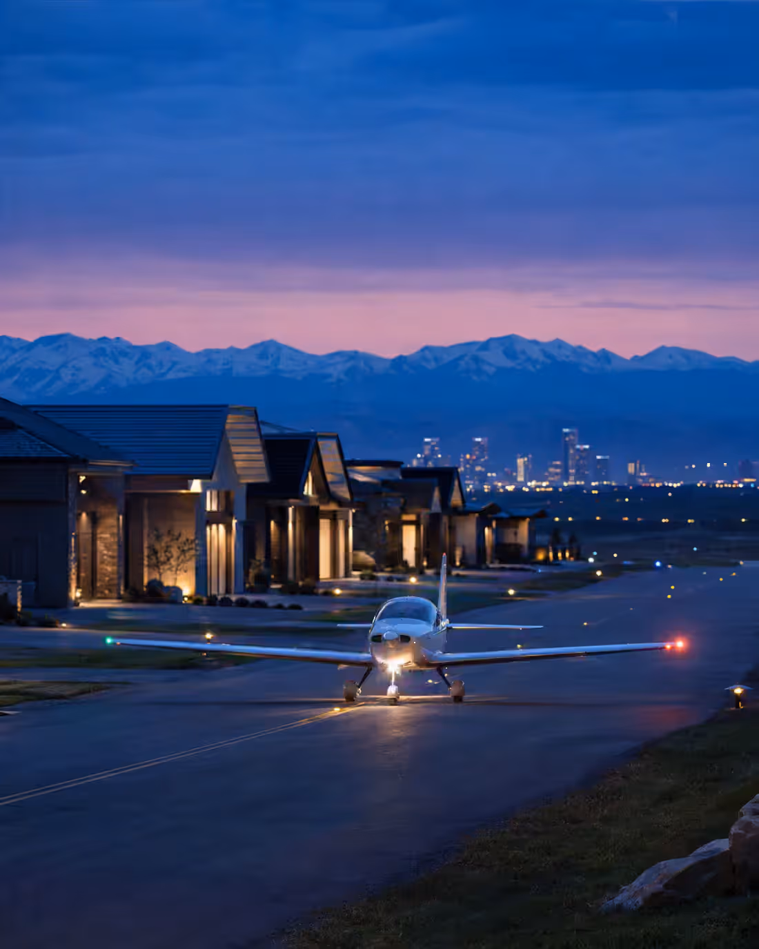 A single-engine aircraft on an airpark taxiway at dusk, landing light on, lit hangar homes to one side and a city skyline beyond