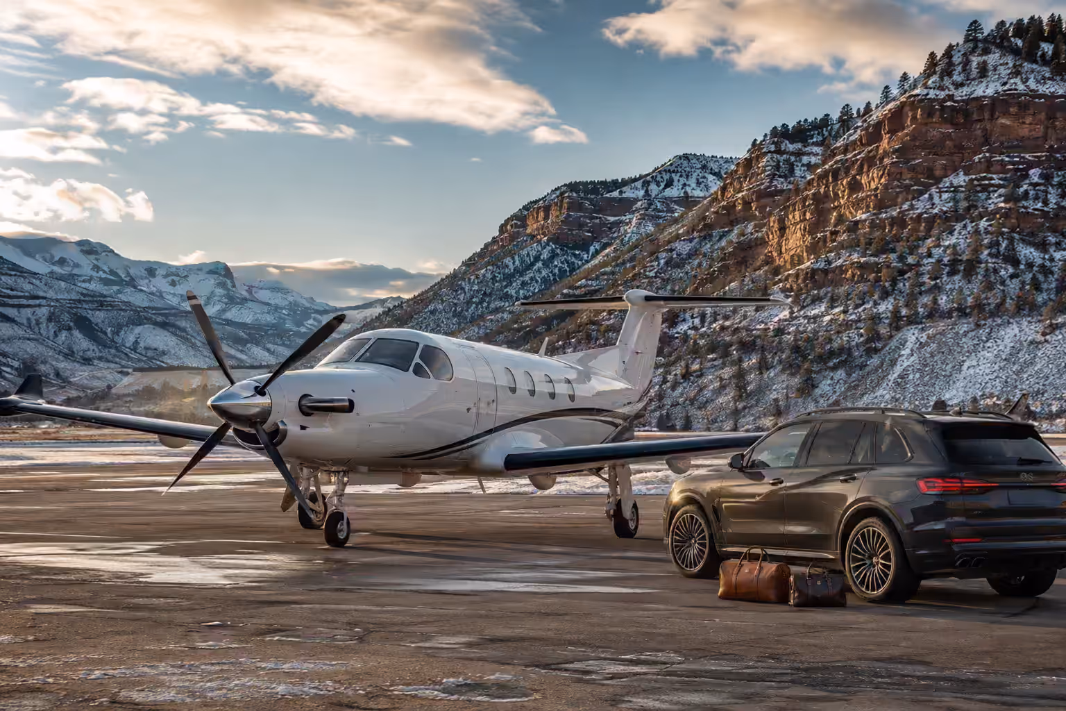 A single-engine turboprop on a winter ramp beside a waiting SUV with two leather holdalls set down, a snow-covered canyon wall behind