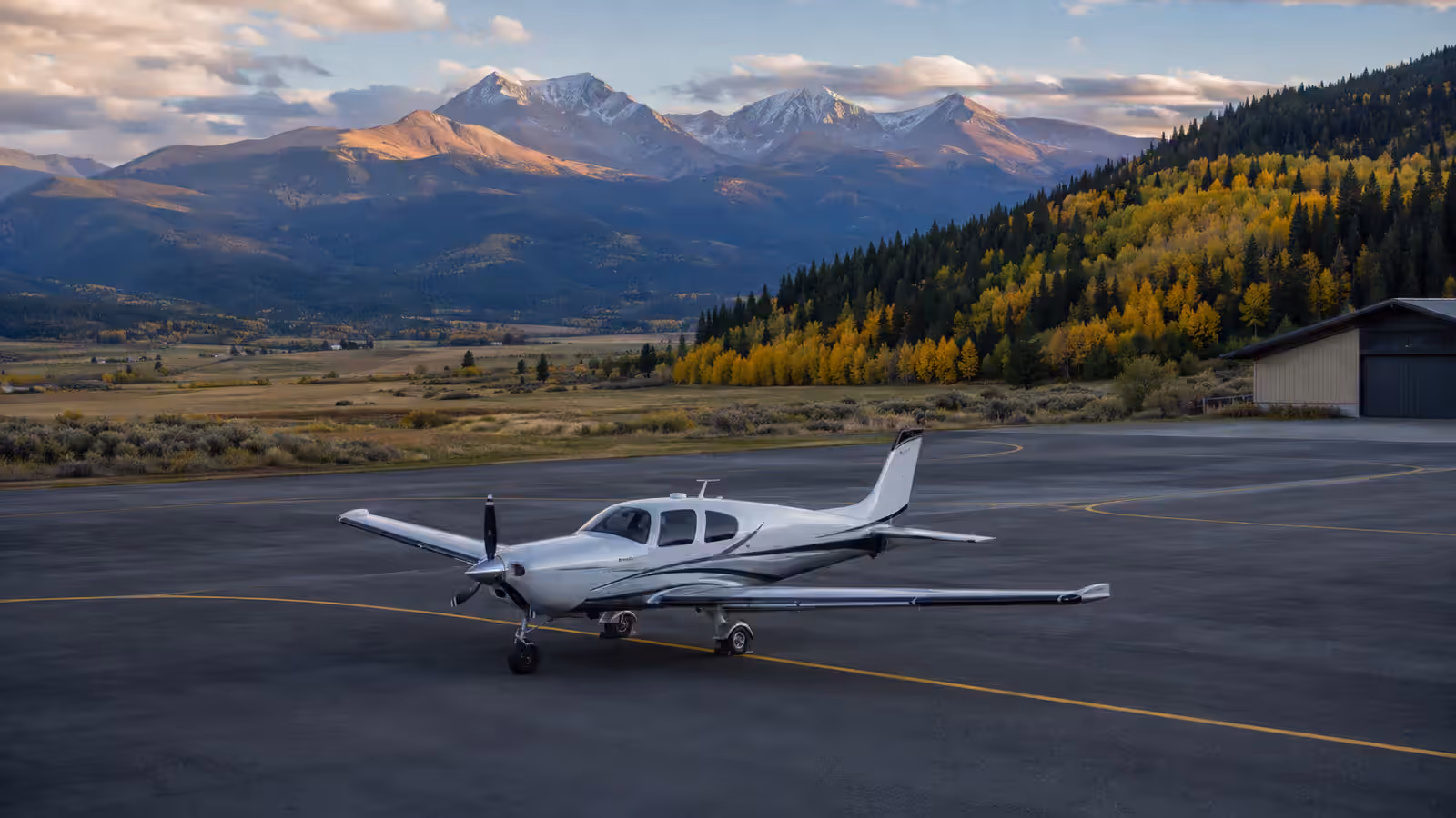 A low-wing single on a valley ramp with a hillside of aspens turning behind it, a hangar at the right and snow-capped peaks across the valley