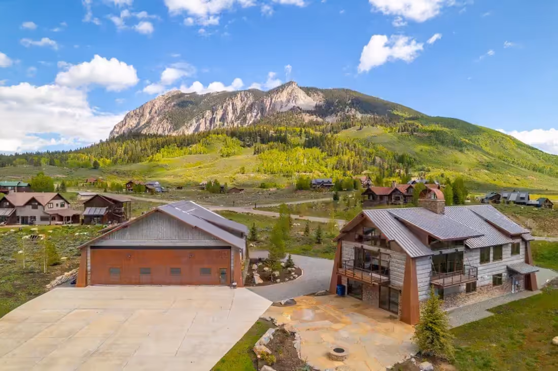 Aerial view of a single parcel carrying two separate buildings, a metal-clad hangar with a broad concrete apron and a timber and stone residence, with a green valley and a rock face behind