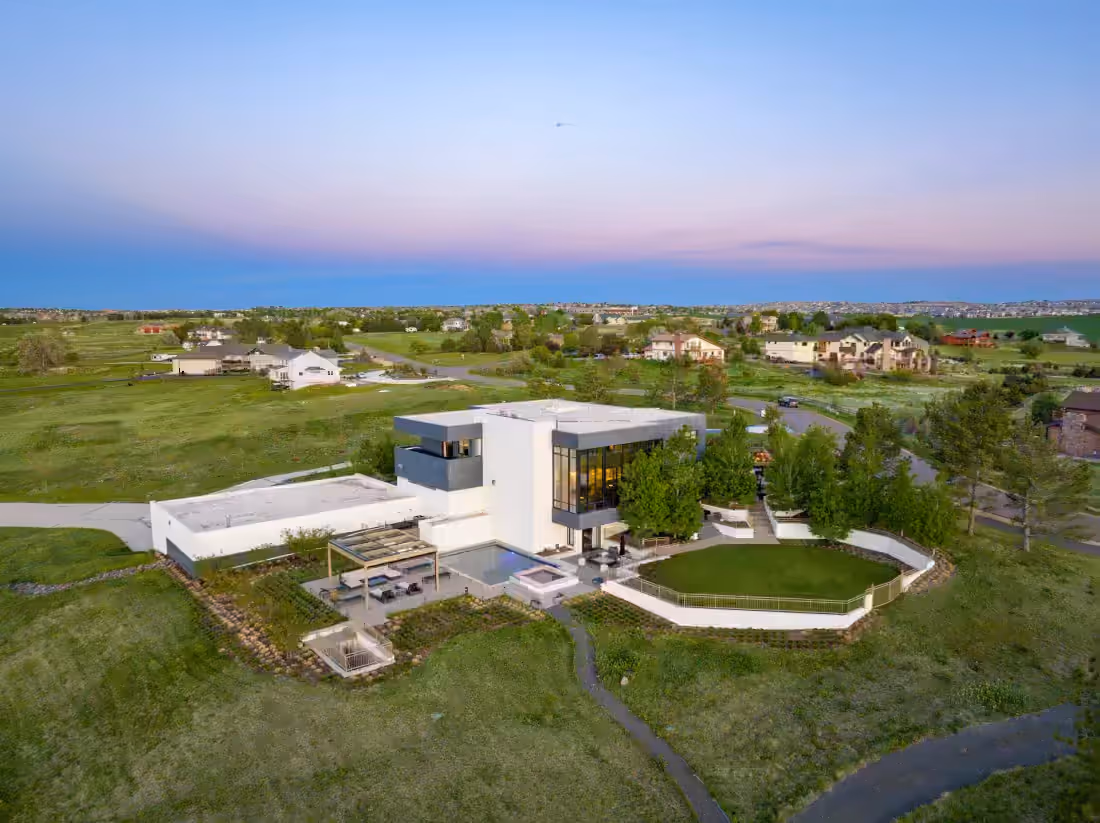 Aerial view at dusk of a modern estate with pool, lawn, and outbuilding on open ground at the edge of a subdivision