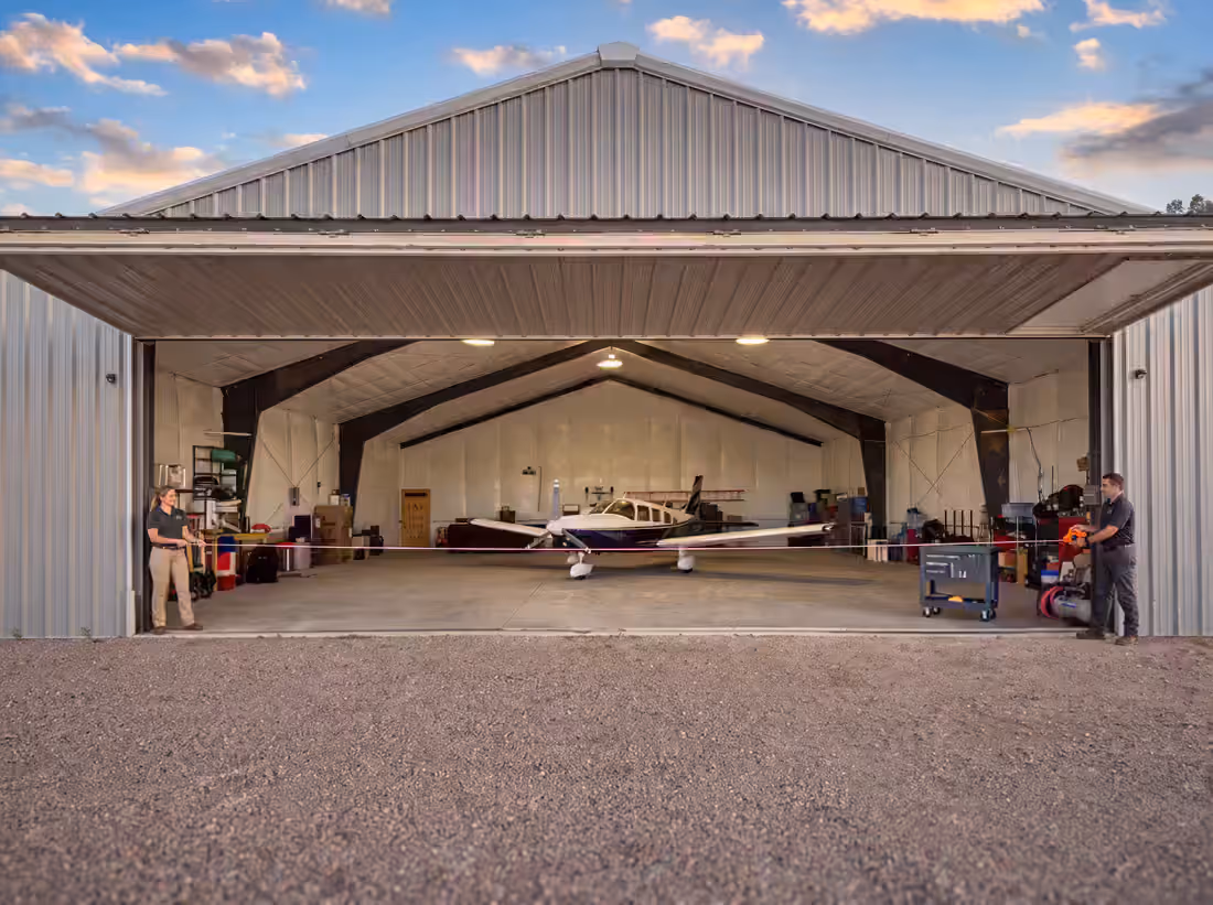 Two people holding a measuring tape across the full width of an open hangar door, a light aircraft inside