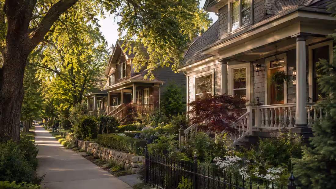 A shaded residential sidewalk running past deep porches, iron railings and planted front gardens