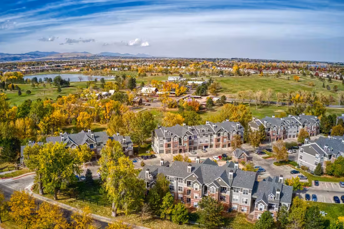 Aerial over Gunbarrel at the turn of autumn: townhomes and low apartment blocks along a golf course, cottonwoods in colour, the Front Range on the horizon