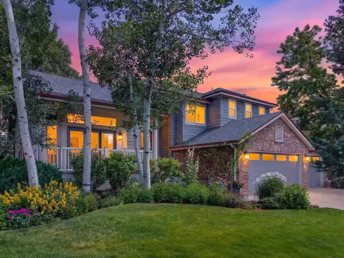 Two-story shingled and brick residence at dusk among aspens, windows lit, planted beds along the drive