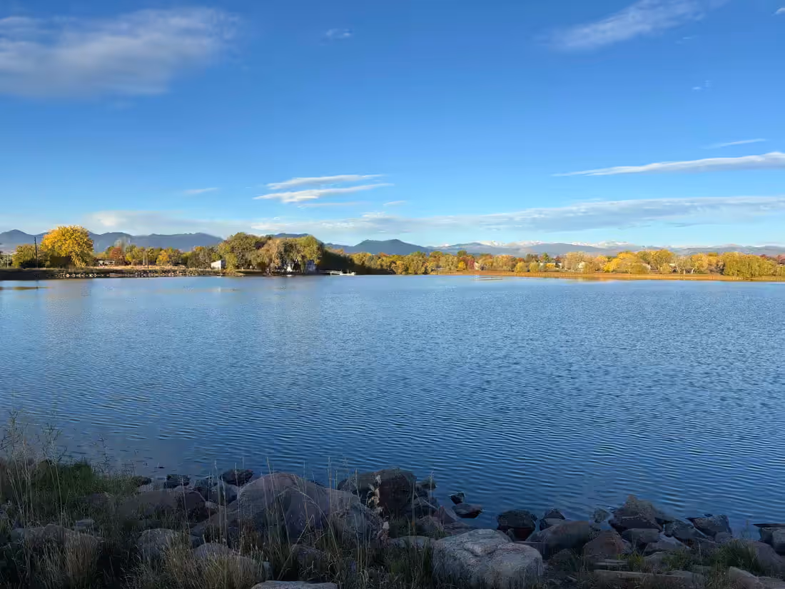 A Front Range reservoir at midday, rocky shore in the foreground, cottonwoods along the far bank and the foothills beyond.