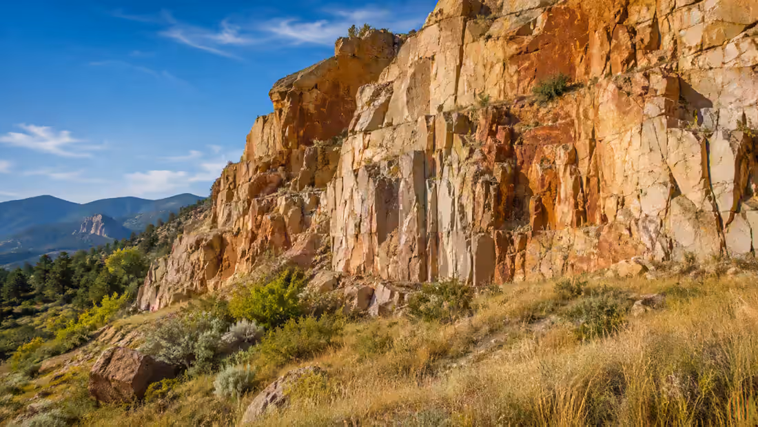 A cut sandstone face in gold and rust above grass and scrub, forested hills falling away beyond it