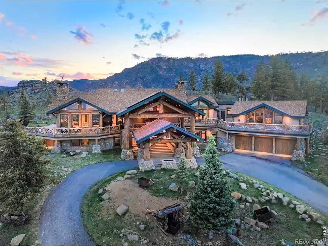A log and stone mountain residence at dusk, rock outcrops rising behind it