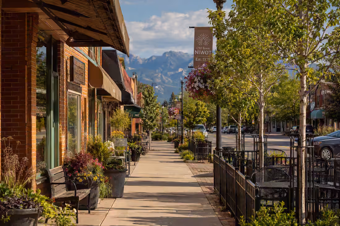 A brick main street under awnings and hanging baskets, planters and iron railings along a swept sidewalk, mountains closing the view.