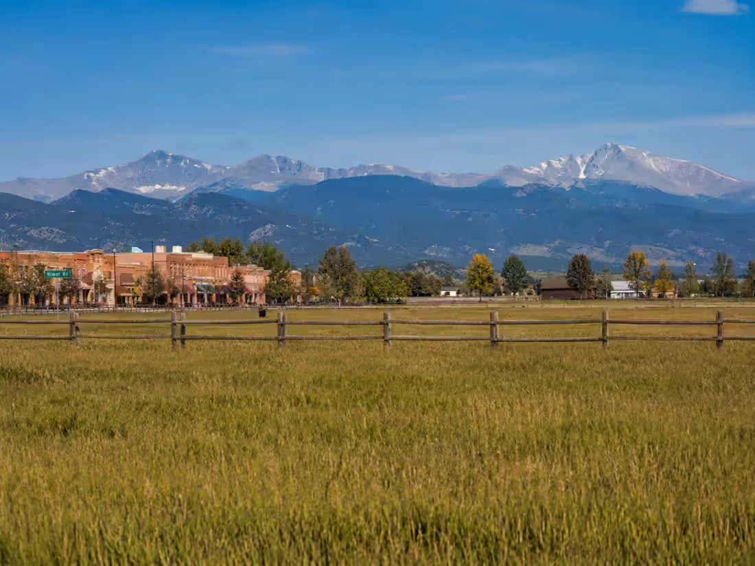 A fenced hay meadow in front of a small brick main street, with the snow-covered high peaks behind it