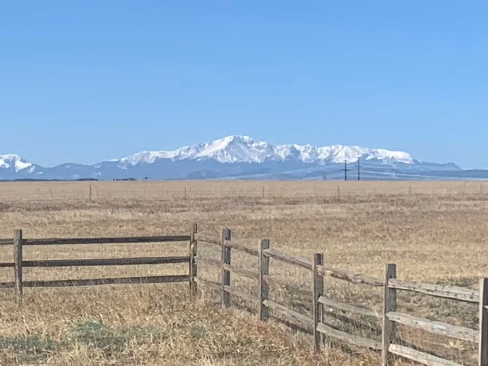 Snow-capped Pikes Peak beyond a split-rail fence and dry prairie grass near Kelly Air Park.
