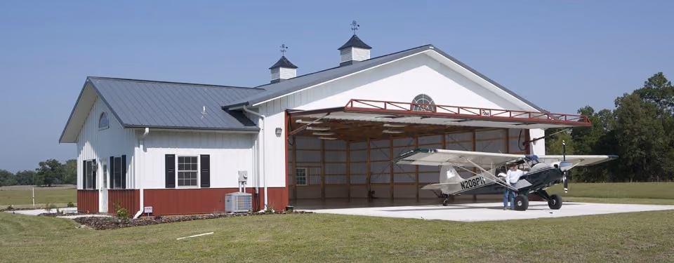 A white pole barn hangar with the hydraulic door open and a taildragger on the pad.