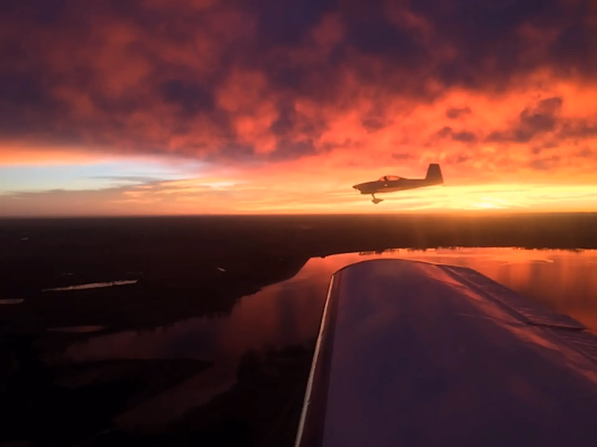 A light aircraft silhouetted against a red sunset, photographed from a second aircraft over water.