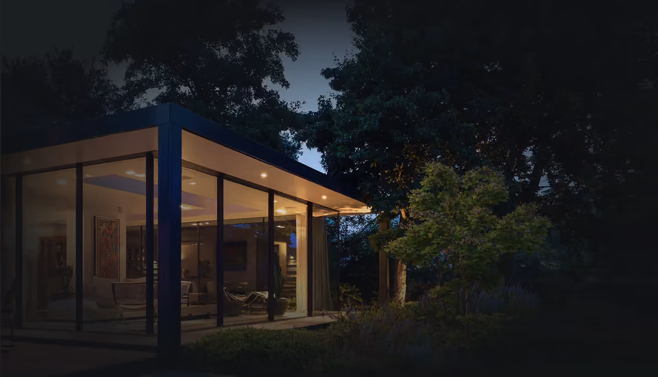 A glass-walled home lit from within at dusk, framed by mature trees